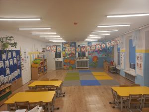 Sala grupy kotki. 3 stoliki żółte, tablica niebieska z materiałami edukacyjnymi.